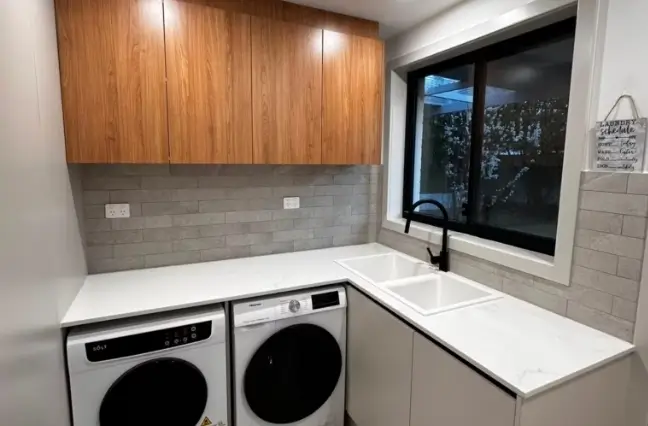 Renovated laundry room featuring modern white subway tiles, custom wood cabinets, and built-in washer dryer setup.